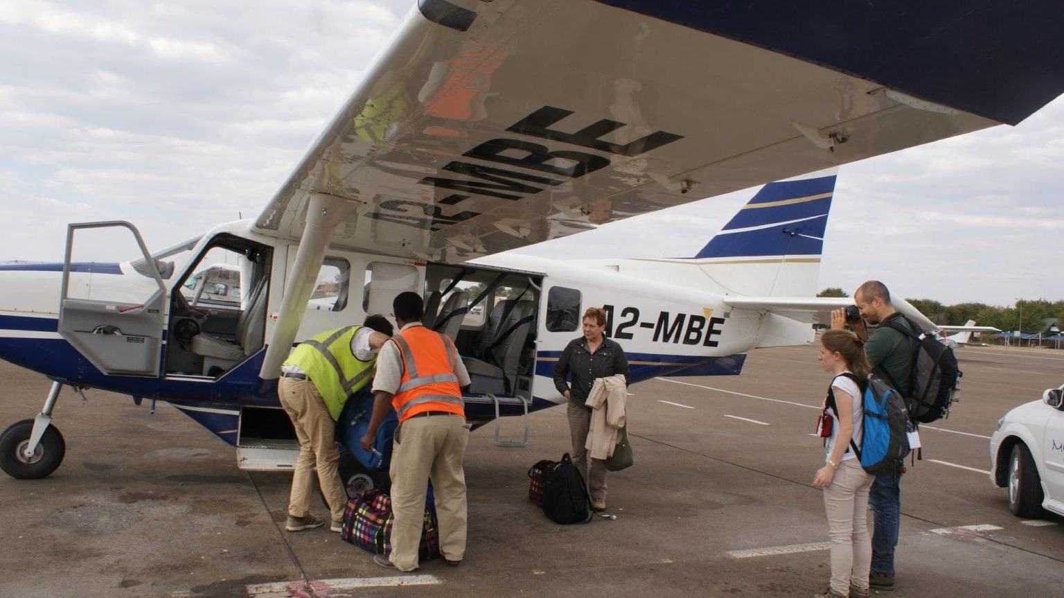 Safari passengers and a bush pilot standing beside a small Cessna on a dirt airstrip in Botswana
