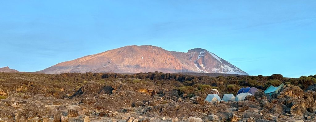 Tents of a high camp on rocky ground below the sunlit summit dome of Kilimanjaro