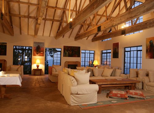 The lounge at Clouds Mountain Gorilla Lodge with sofas, a stone fireplace and tall windows at dusk