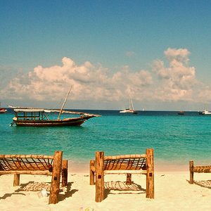 Wooden sun loungers on white sand facing a turquoise sea with a dhow moored offshore