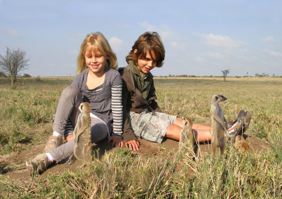 Two children in neutral clothes and trail shoes sitting on open grassland beside a group of meerkats