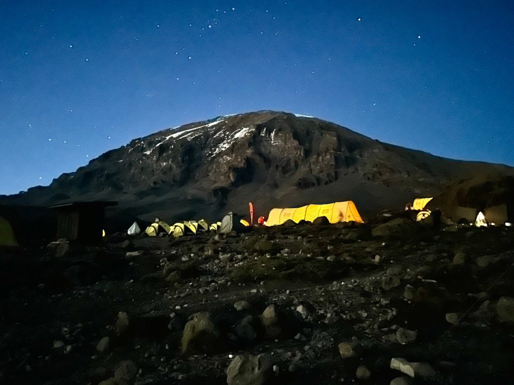 Lit tents of a Kilimanjaro camp at night below the dark dome of Kibo