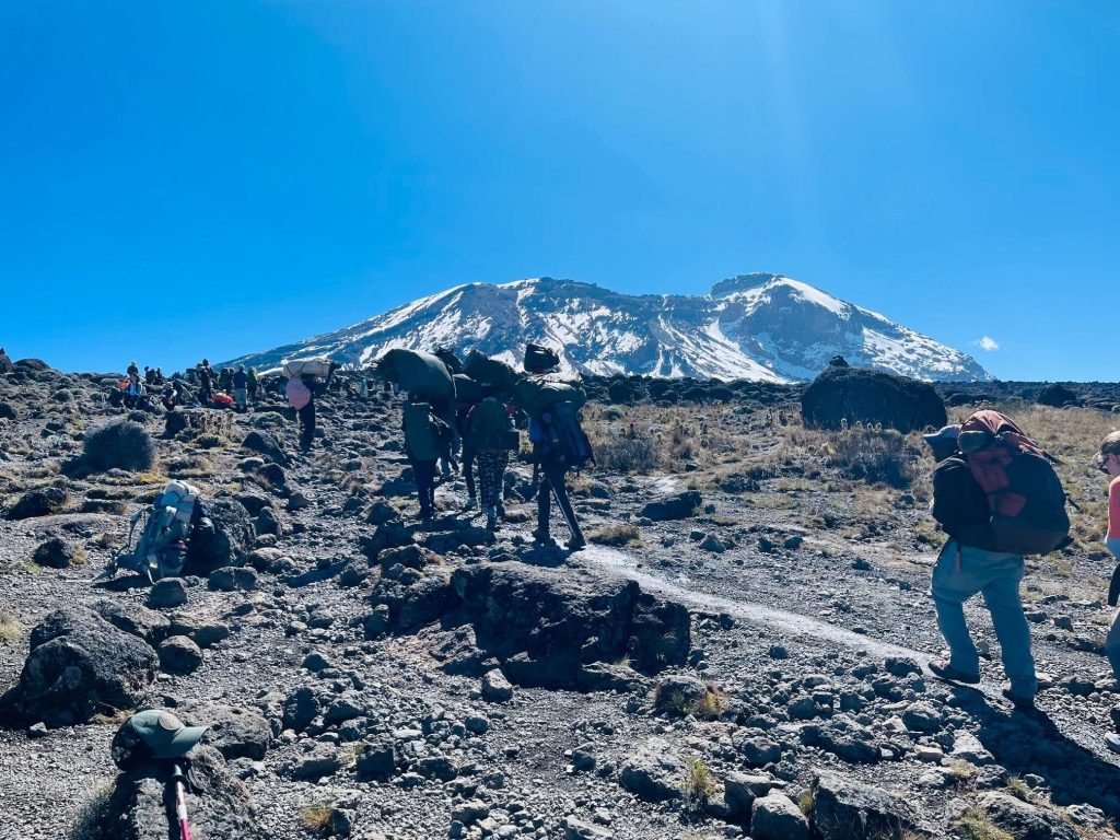 Porters carrying duffel bags and trekkers on a rocky trail toward the snow capped Kibo summit of Kilimanjaro