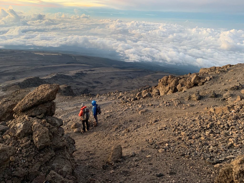 Two trekkers crossing bare alpine desert scree high on Kilimanjaro above a sea of cloud