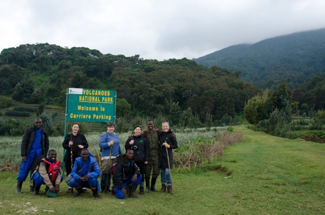 Gorilla trekkers and porters at the Volcanoes National Park trailhead sign in Rwanda, wearing long trousers tucked into boots